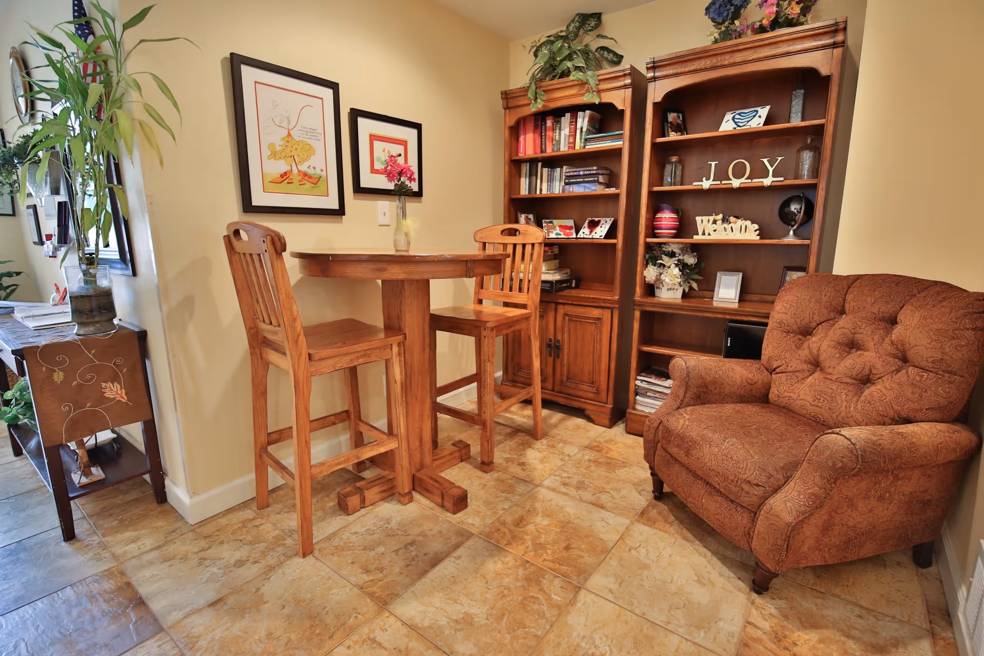 A cozy corner in a living area featuring a wooden high table with two matching high chairs, a large cushioned armchair with a patterned fabric, and two wooden bookshelves filled with books and decorative items including a sign that says 'JOY' and 'Welcome'. The floor is tiled and the walls are painted a warm beige color. There are framed pictures on the wall and a small vase with flowers on the table.