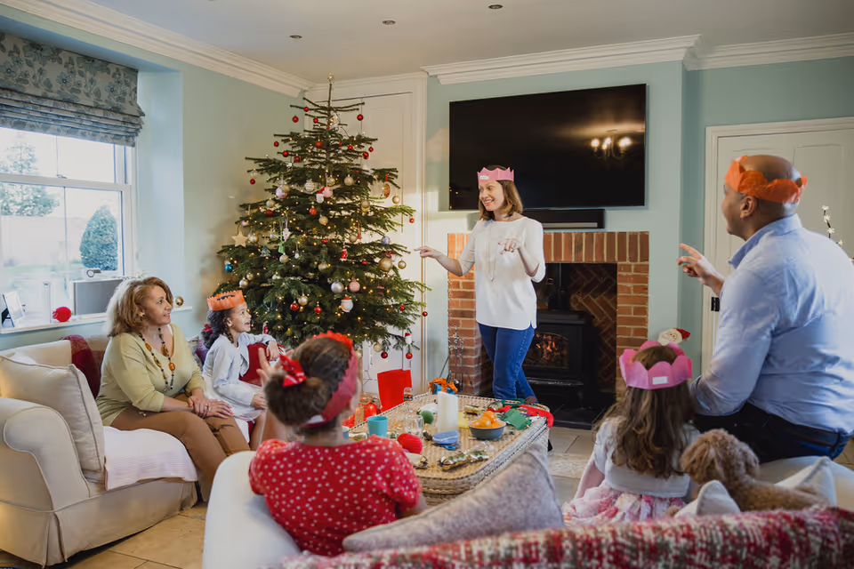 A family gathered in a living room decorated for Christmas with a Christmas tree, presents, and festive decorations. Several people, including children and adults, are wearing colorful paper crowns and appear to be enjoying a holiday celebration. The room has light blue walls, a large window, a fireplace with a TV mounted above it, and comfortable seating.