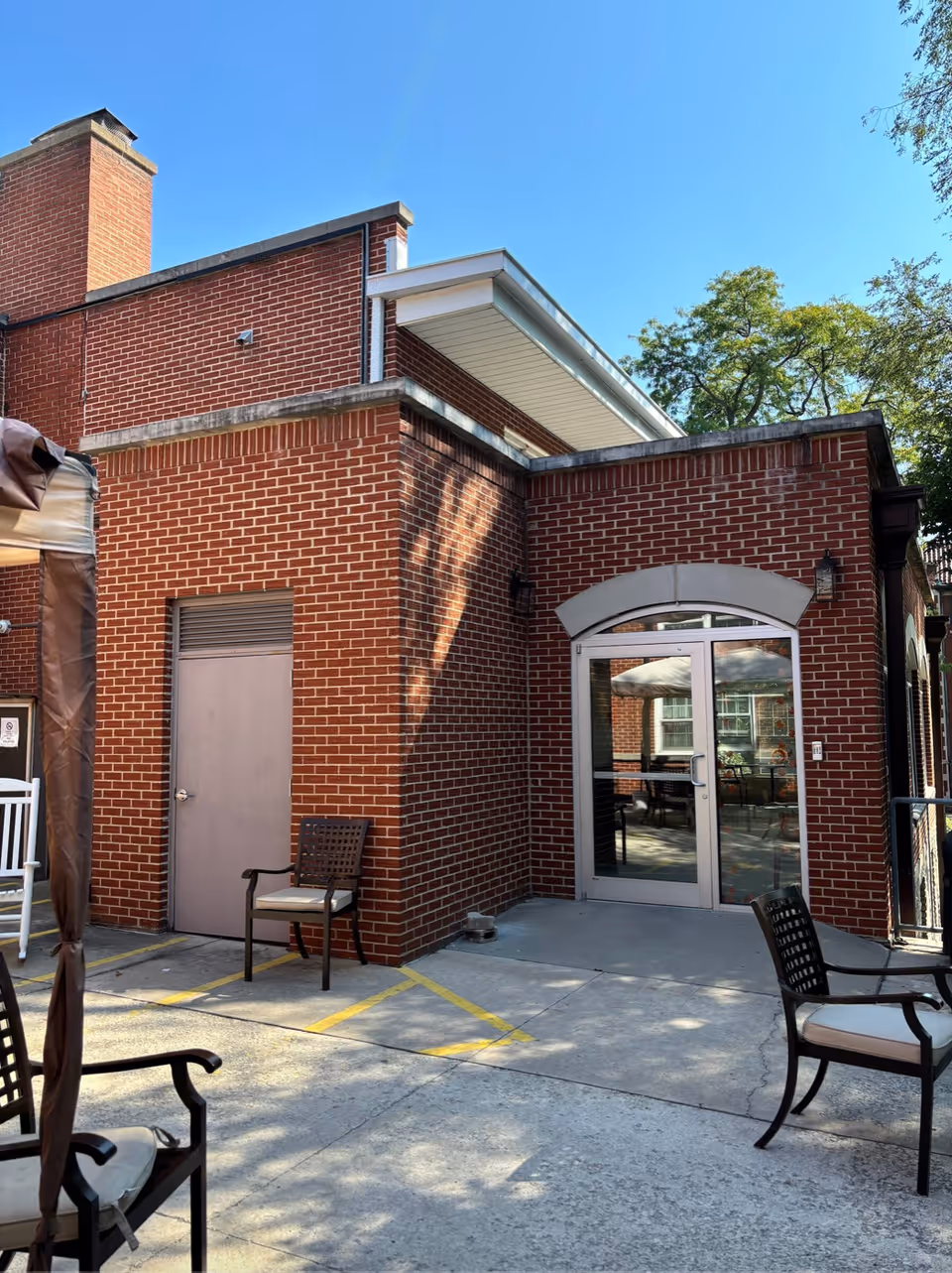 Brick building exterior with a small concrete patio, metal chairs, and glass double doors under a clear blue sky.