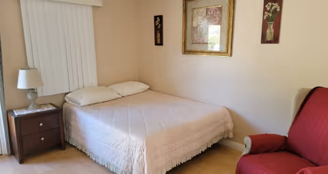 A bedroom in a senior living facility featuring a bed, nightstand, lamp, and red chair.