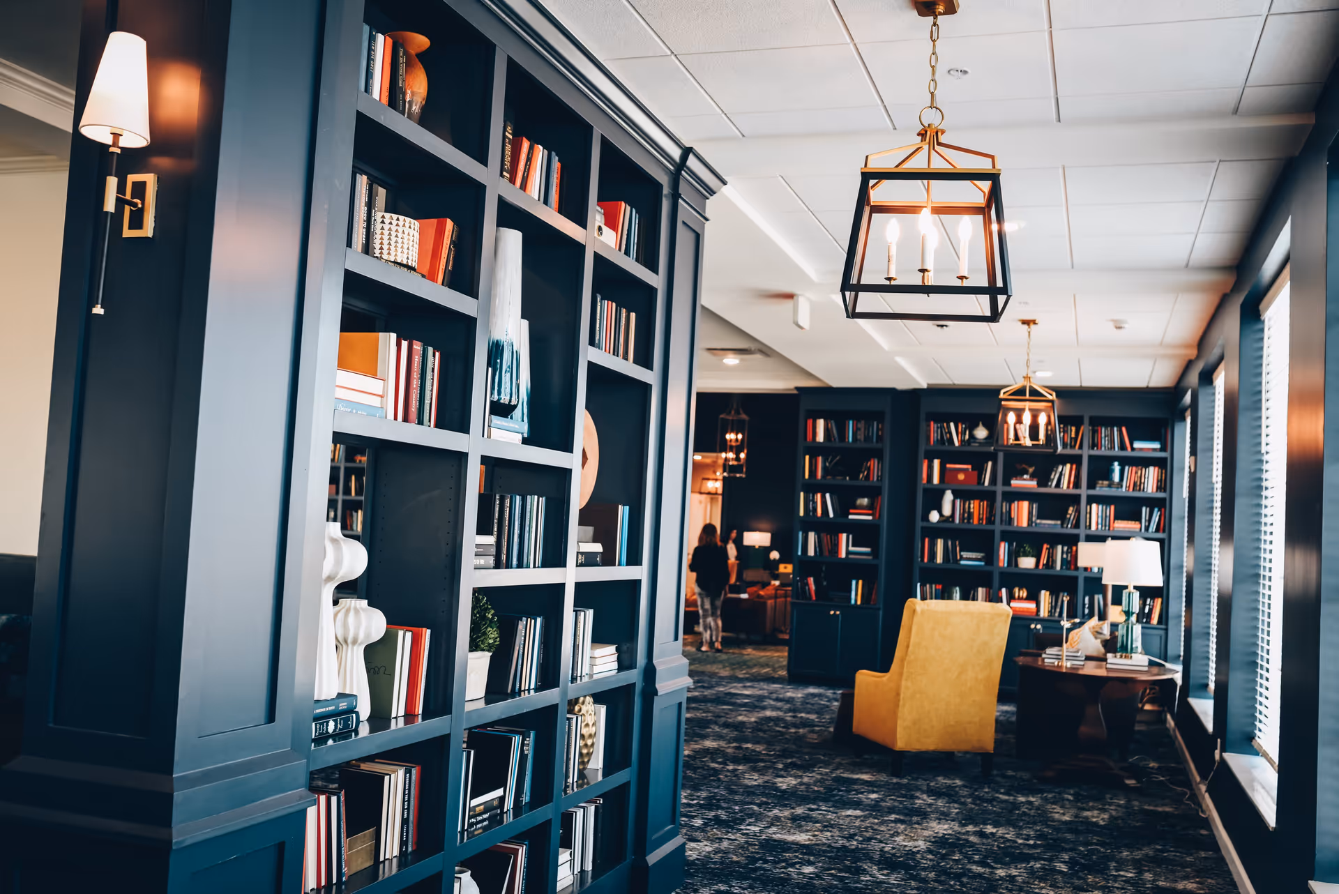 Interior of a cozy library or lounge area with dark blue built-in bookshelves filled with books and decorative items. The room features a yellow armchair, a wooden side table with a lamp, large windows with blinds, and hanging pendant lights with candle-style bulbs. A person is seen walking in the background.
