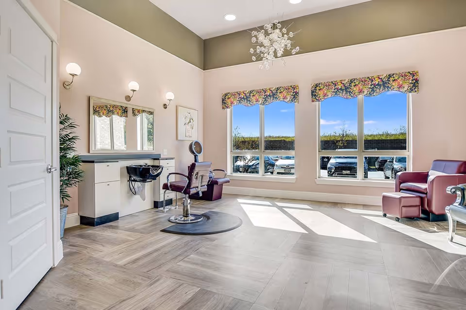 A bright and spacious room with large windows letting in natural light, featuring a salon chair and hair washing station on the left, a maroon leather armchair on the right, floral valances above the windows, and a modern chandelier hanging from the ceiling.