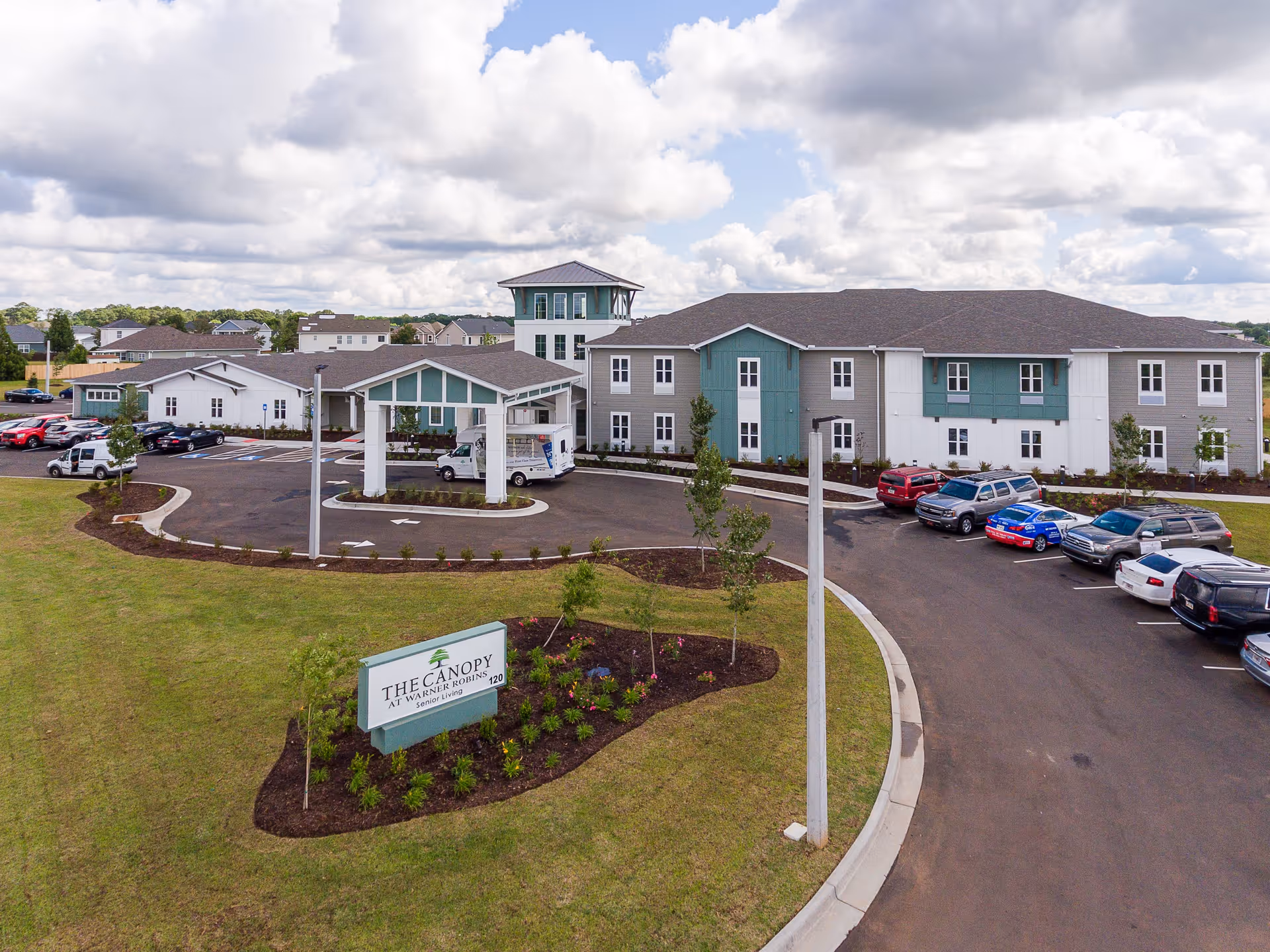 Exterior view of The Canopy at Warner Robins senior living facility showing a large two-story building with a covered entrance, multiple parked cars, landscaped grounds, and a sign with the facility name in front.