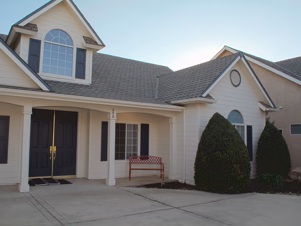 Front exterior of a light-colored residential building with double black doors, a porch with a red bench, and neatly trimmed bushes.
