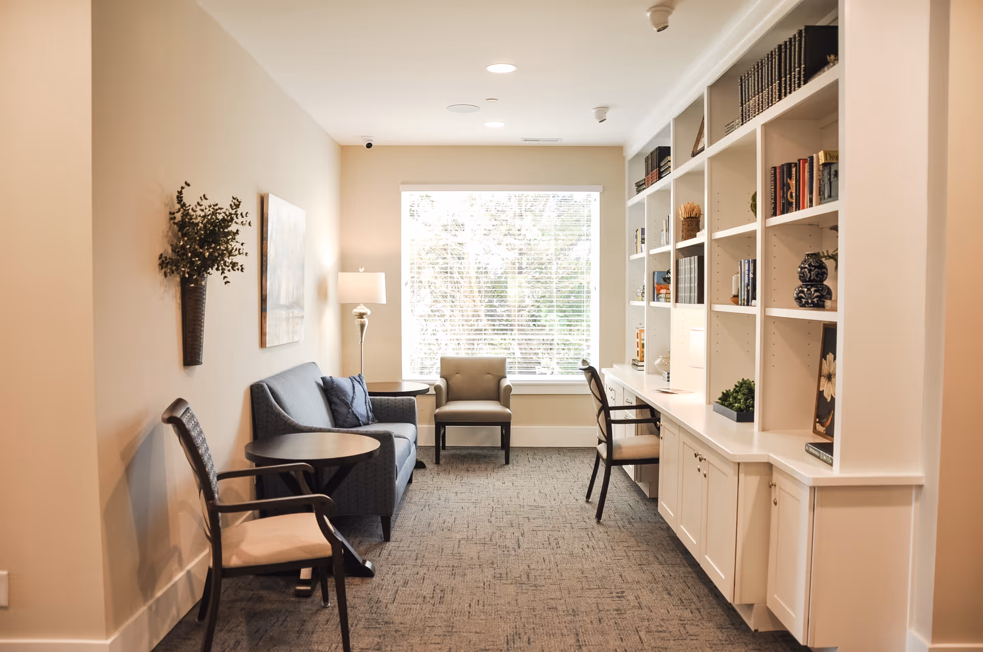 A cozy interior room with a large window letting in natural light. The room features a blue sofa with two cushions, a beige armchair, a round side table, and a floor lamp. On the opposite wall, there is a built-in white bookshelf filled with books and decorative items, along with a desk area and chairs. The walls are painted a light beige color, and the floor is carpeted in a neutral tone.