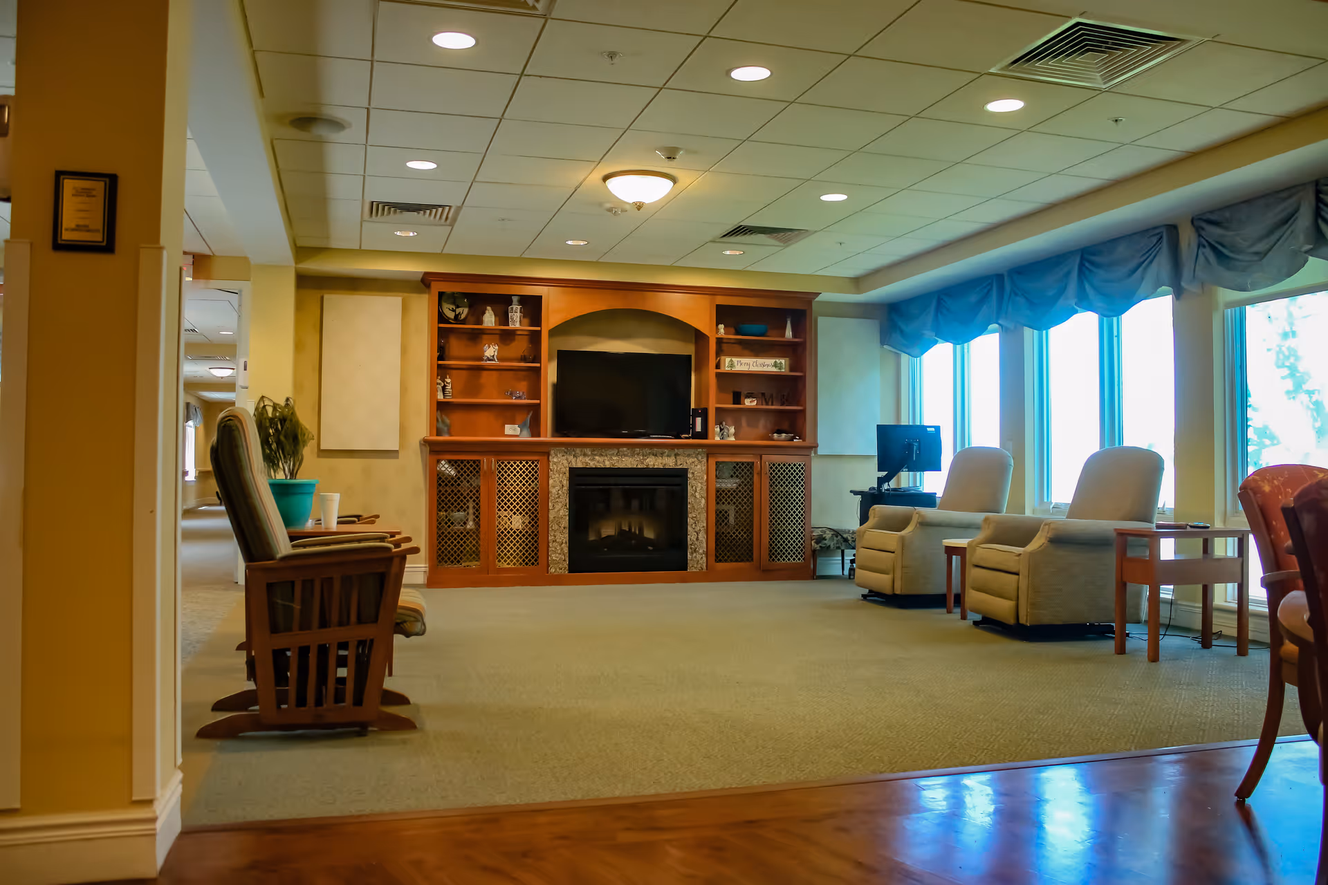A cozy living room area in a senior living facility with a wooden entertainment center featuring a TV and a fireplace. The room has several comfortable chairs, large windows with blue valances letting in natural light, and a carpeted floor with some wooden flooring in the foreground.