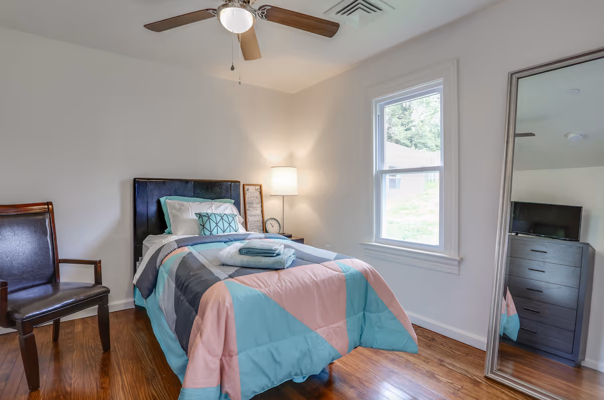 Single-occupancy bedroom with a twin bed dressed in colorful geometric bedding, a bedside lamp, chair, window, ceiling fan, and a large floor mirror reflecting a dresser.