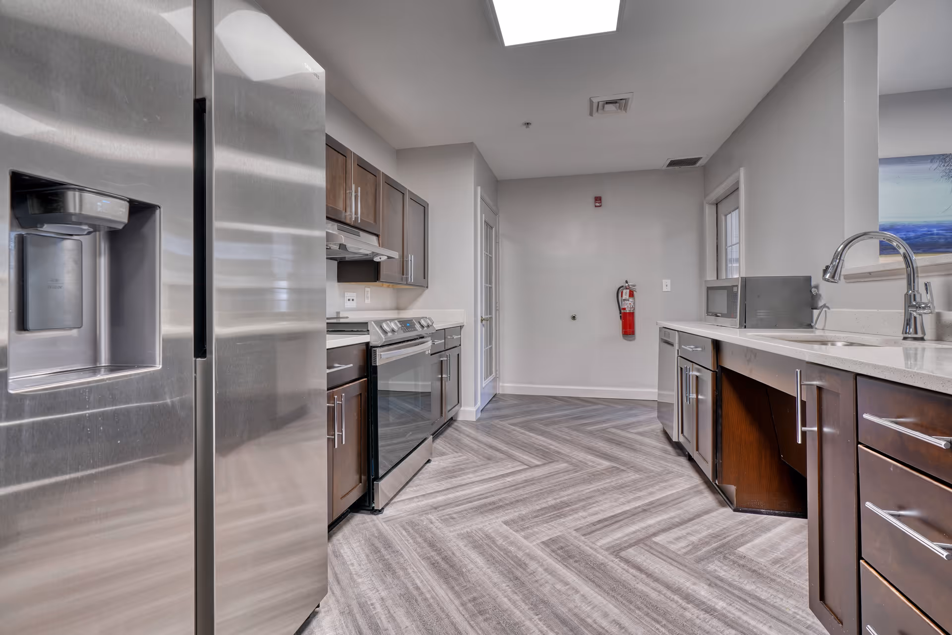 Modern communal kitchen with stainless steel appliances, dark wood cabinets, a sink, and chevron-patterned flooring.