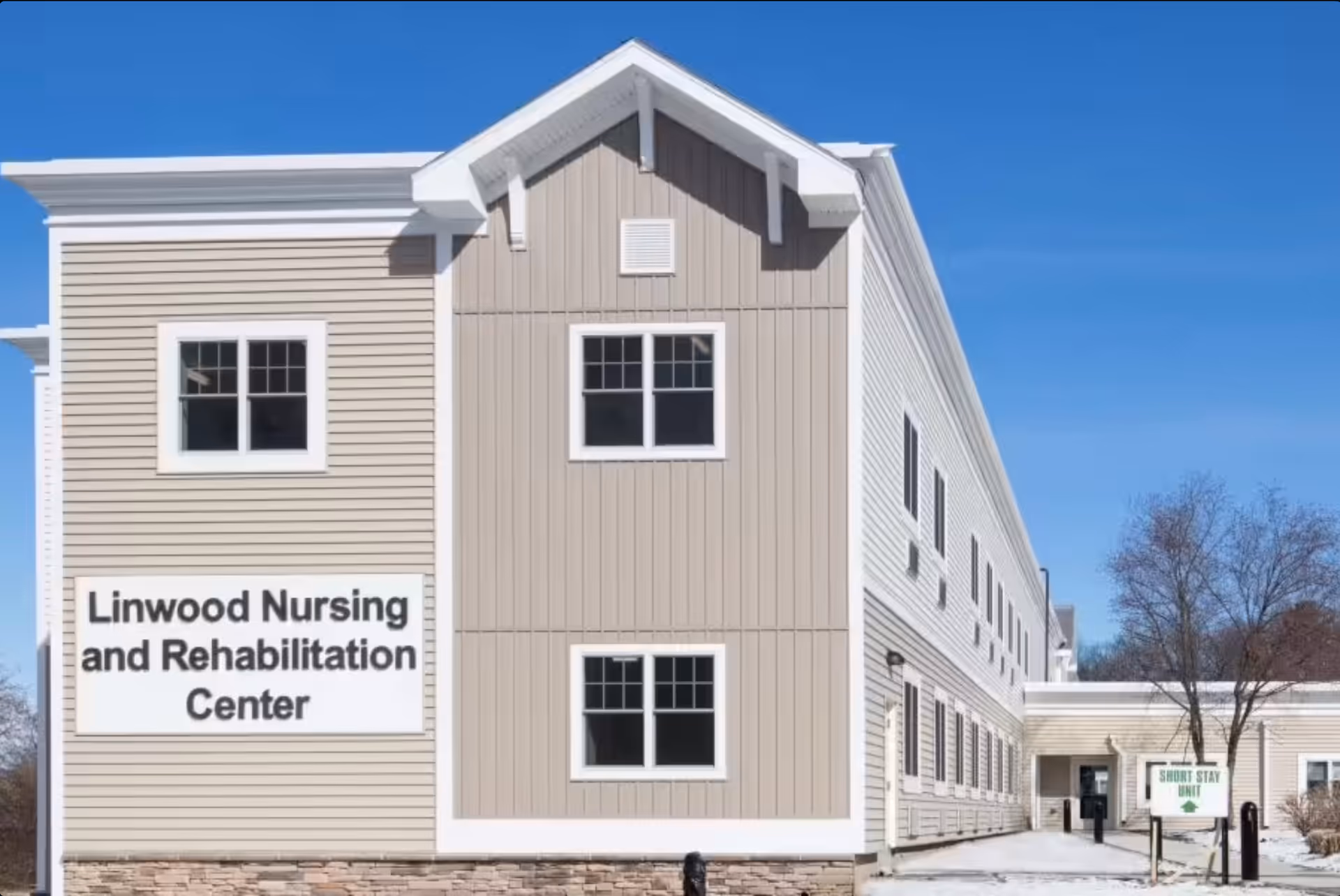 Exterior view of Linwood Nursing and Rehabilitation Center building with beige and white siding, multiple windows, and a sign indicating the facility's name. There is a smaller sign pointing to the Short Stay Unit near the entrance. The sky is clear and blue.