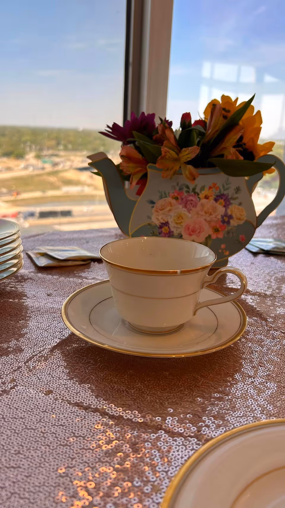 A china teacup and saucer on a sequined tablecloth with a floral teapot centerpiece and a window view in the background.