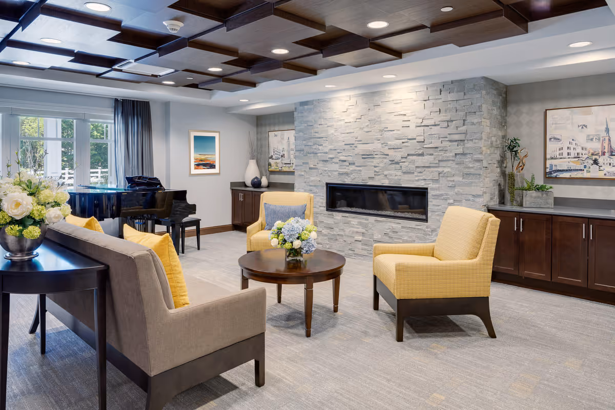 A cozy living room area with a modern stone fireplace, two yellow armchairs, a gray sofa with yellow pillows, a round wooden coffee table with a flower arrangement, and a black grand piano near a window with gray curtains. The room has a coffered ceiling with recessed lighting and artwork on the walls.