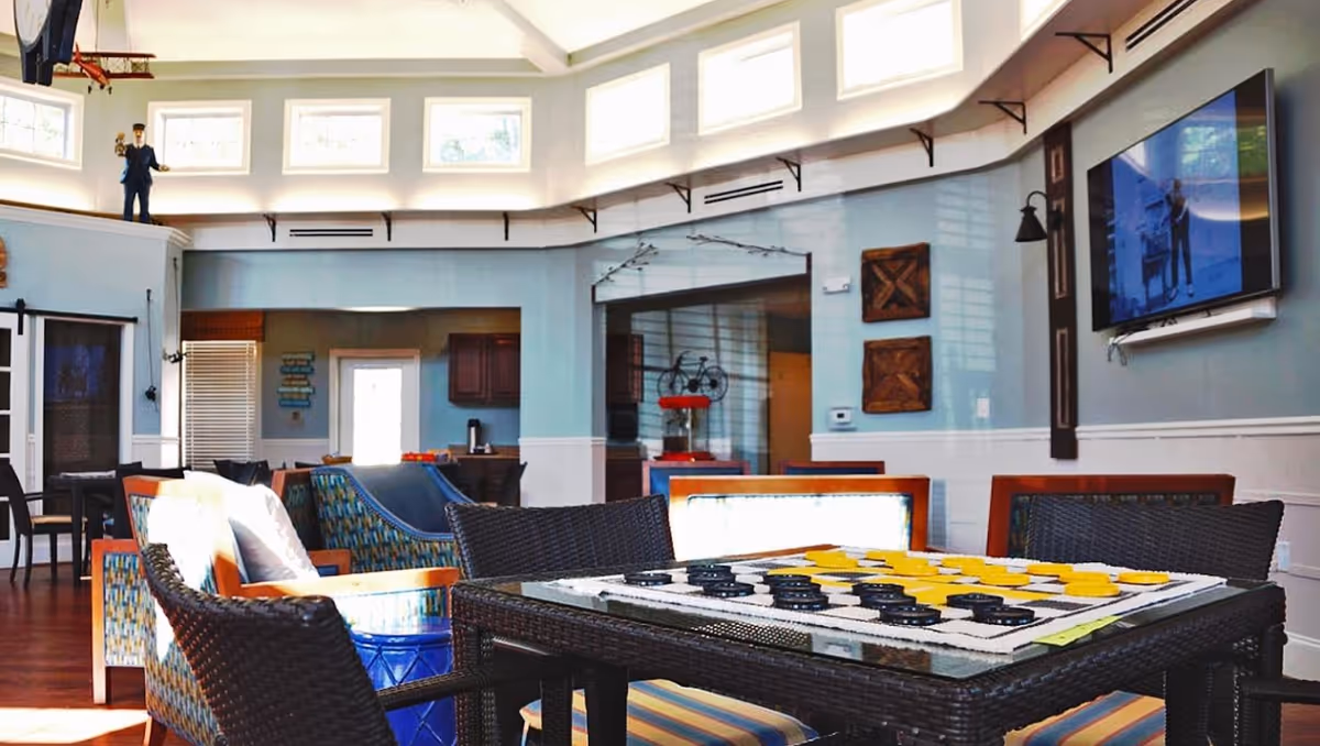 Bright common lounge with wicker chairs and a table set up with a board game, seating area, and a wall-mounted TV.