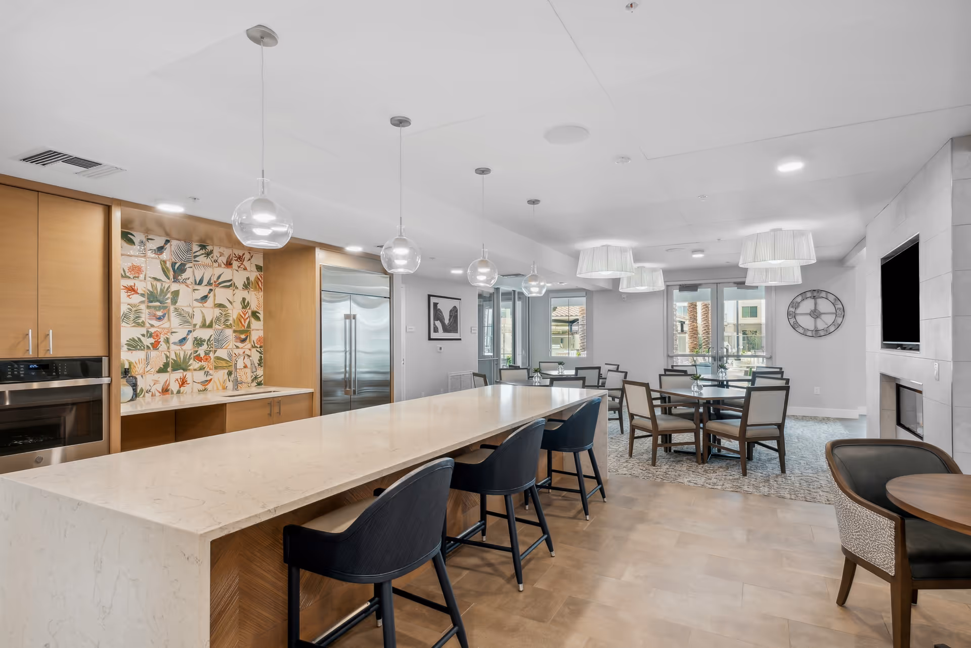 Bright communal kitchen and dining area with a long marble island, pendant lights, bar stools, and multiple dining tables.
