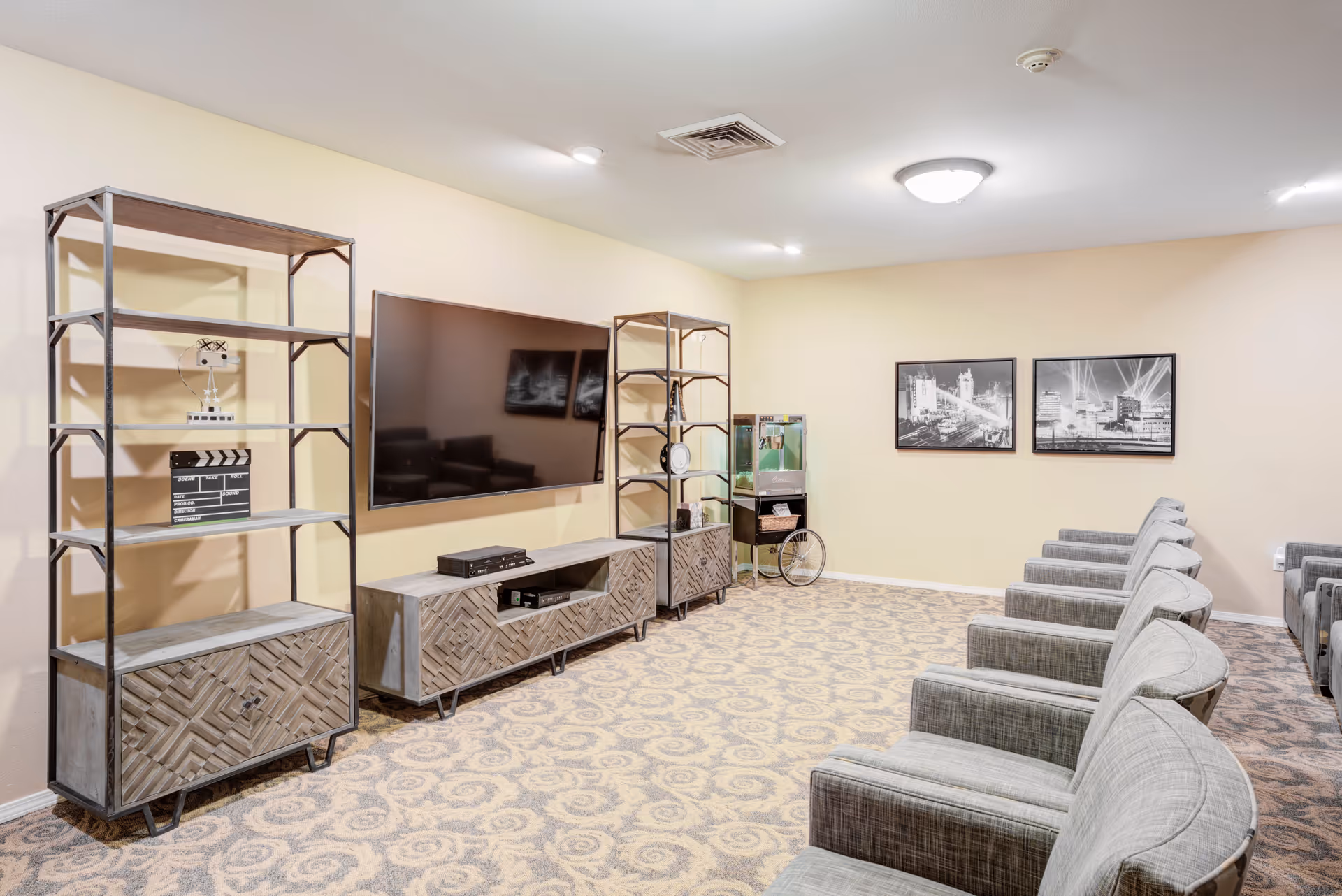 A senior living facility common room with a large flat-screen TV mounted on the wall, flanked by two wooden shelving units. Several gray upholstered armchairs are arranged in rows facing the TV. The room has patterned carpet flooring, beige walls, and two black and white framed pictures on the far wall. A popcorn machine on a stand is visible in the corner.