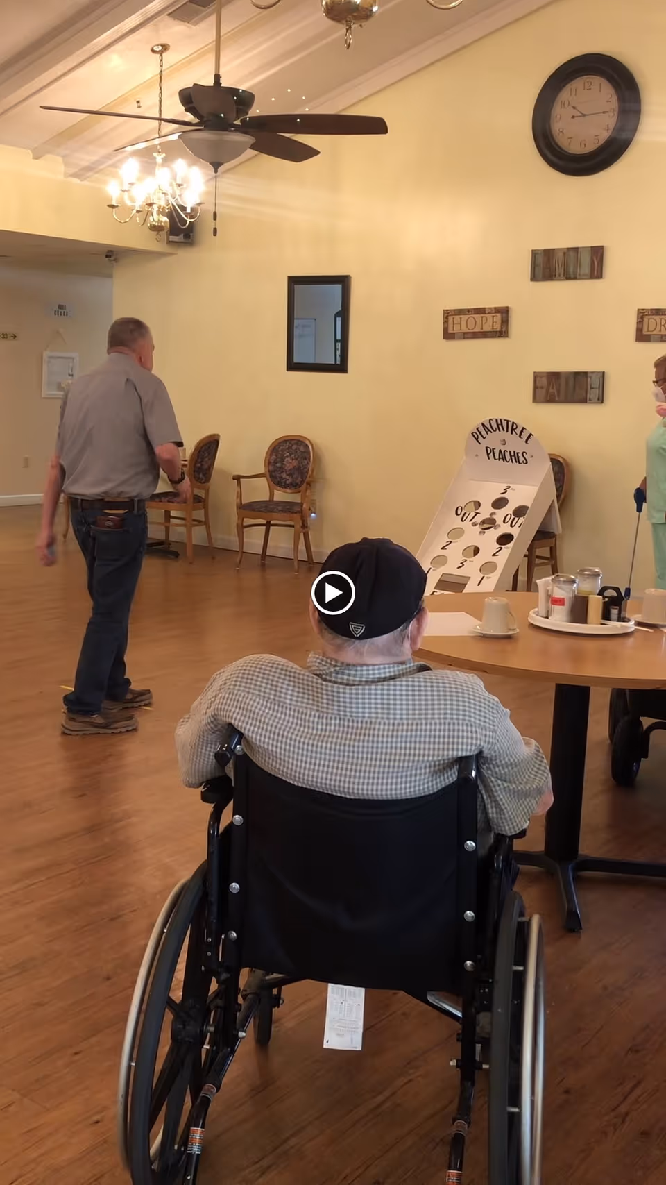 Residents in a senior living facility common room with a person in a wheelchair facing a table, others standing, and a 'Peachtree Peaches' game board against the wall.