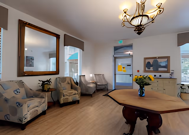 Sunlit lobby with armchairs along the wall, a central wooden table with a vase of flowers, a chandelier, mirror, and an entrance door.