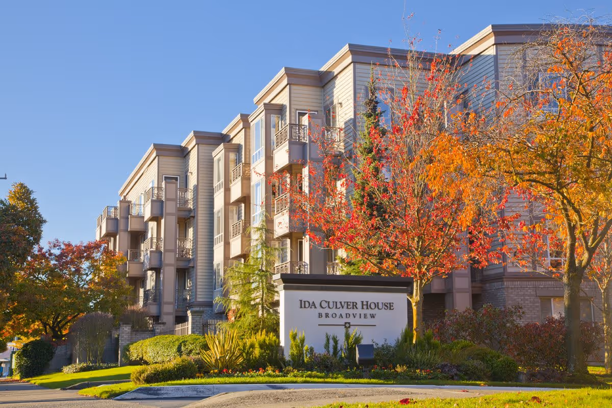 Exterior view of Ida Culver House Broadview, a multi-story senior living facility with balconies, surrounded by trees with autumn foliage under a clear blue sky.