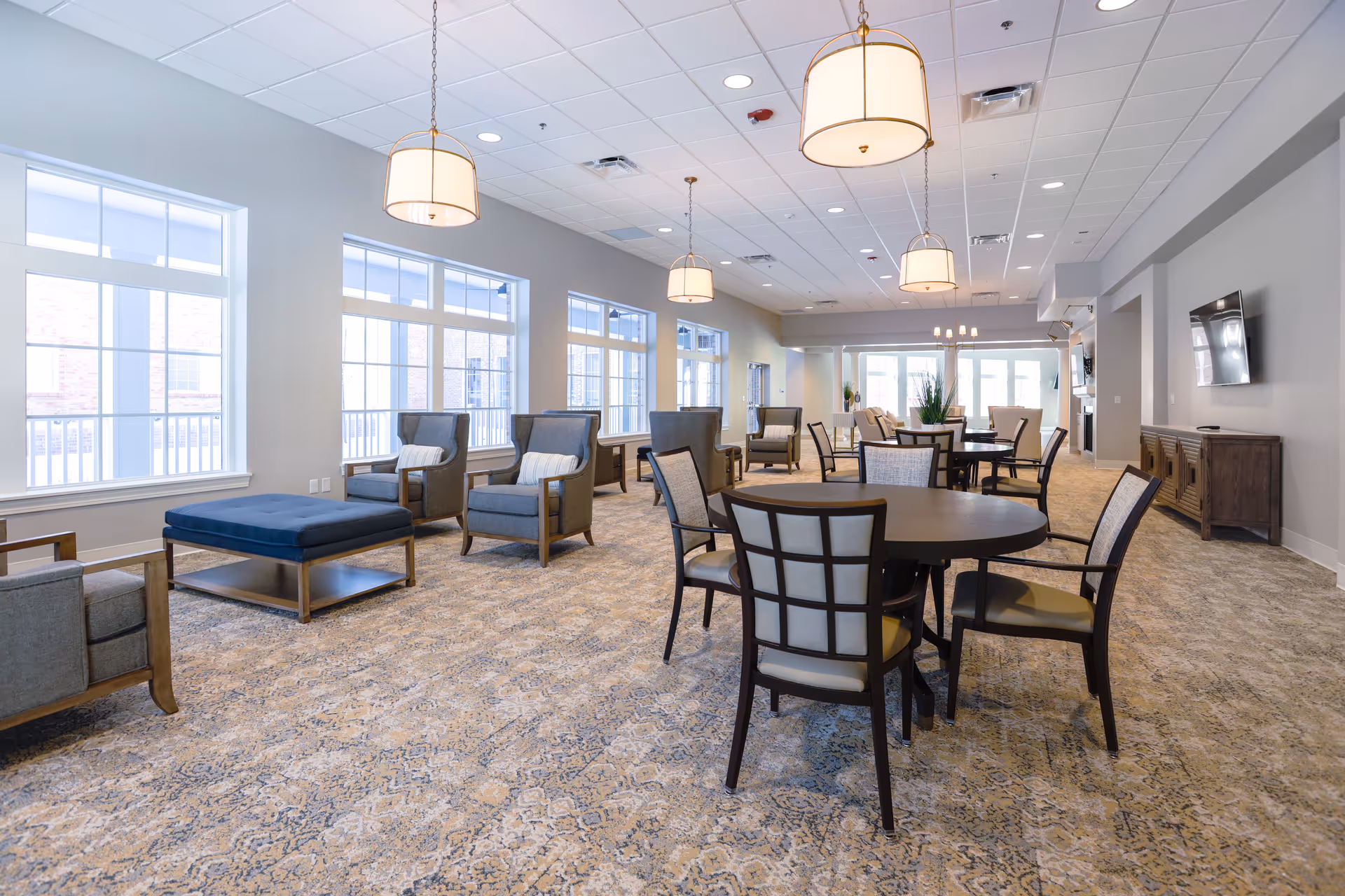 A spacious and well-lit common area in Sayre Christian Village featuring multiple round tables with chairs, several armchairs with cushions, large windows allowing natural light, hanging pendant lights, a wall-mounted TV, and a patterned carpeted floor.