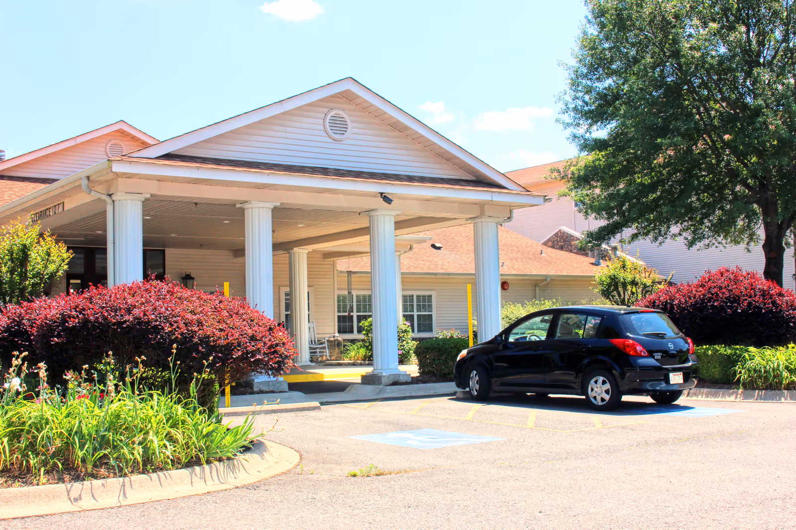 Front entrance of Garrett Woods Senior Living Community with a covered portico supported by white columns, landscaped bushes, and a parked black car.