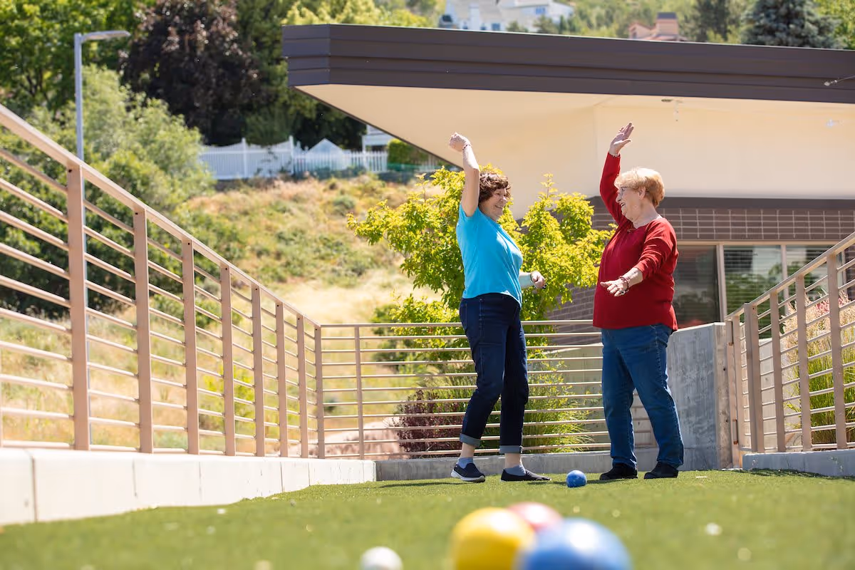 Two women on a grassy outdoor court raise their arms and smile while playing bocce.