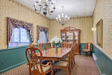 A traditional dining room with a long wooden table surrounded by wooden chairs with cushioned seats. The walls are decorated with floral wallpaper and green wainscoting. Two windows with patterned valances let in natural light. Two chandeliers hang from the ceiling, and a wooden china cabinet filled with dishes is against the far wall. A vase with white flowers is centered on the table.