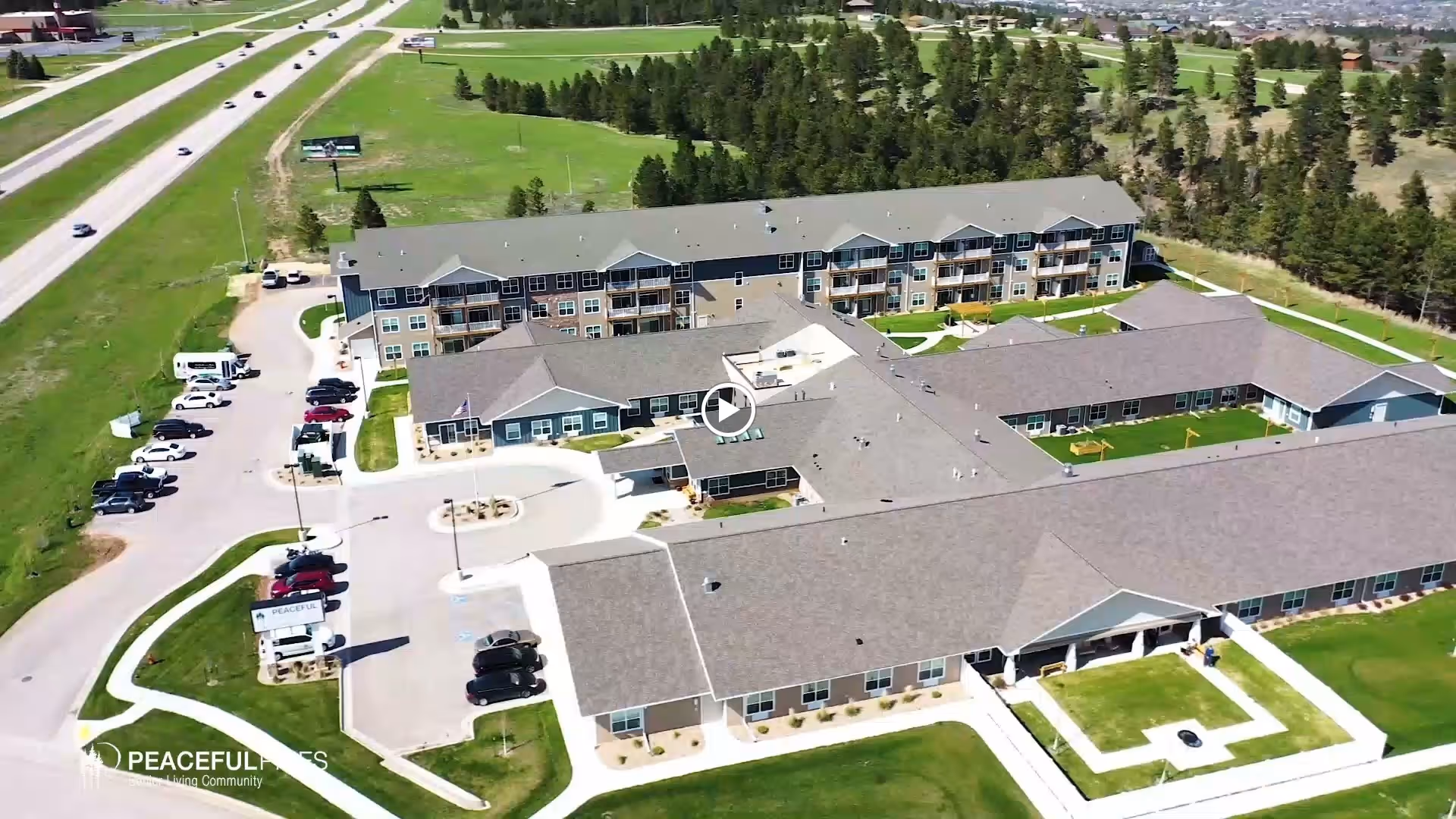 Aerial view of a multi-building senior living community with parking lots, green lawns, and a nearby highway.