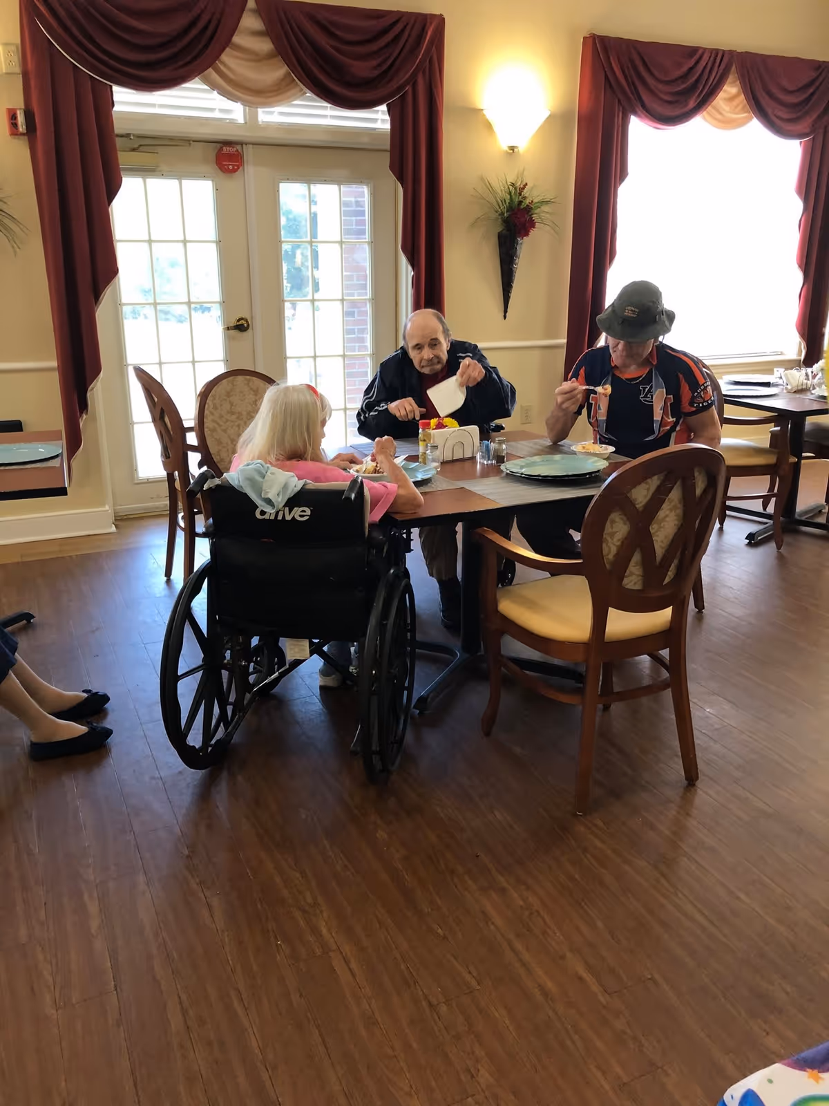 Three elderly individuals sitting around a dining table in a well-lit room with wooden floors and large windows covered with red and beige curtains. One person is in a wheelchair, and they appear to be eating or preparing to eat a meal together.