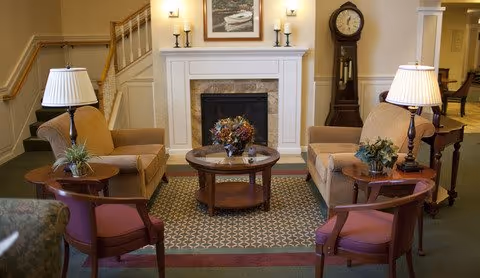 Cozy common room with armchairs and chairs arranged around a fireplace and coffee table, lit by table lamps and featuring a grandfather clock.