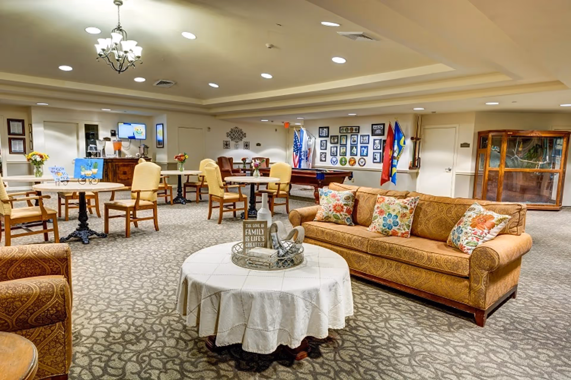 A spacious and well-lit common area in a senior living facility featuring a patterned carpet, a round table with a white tablecloth and decorative items, a brown upholstered sofa with colorful floral pillows, several round tables with chairs, a pool table, and a wall decorated with framed photos and flags.