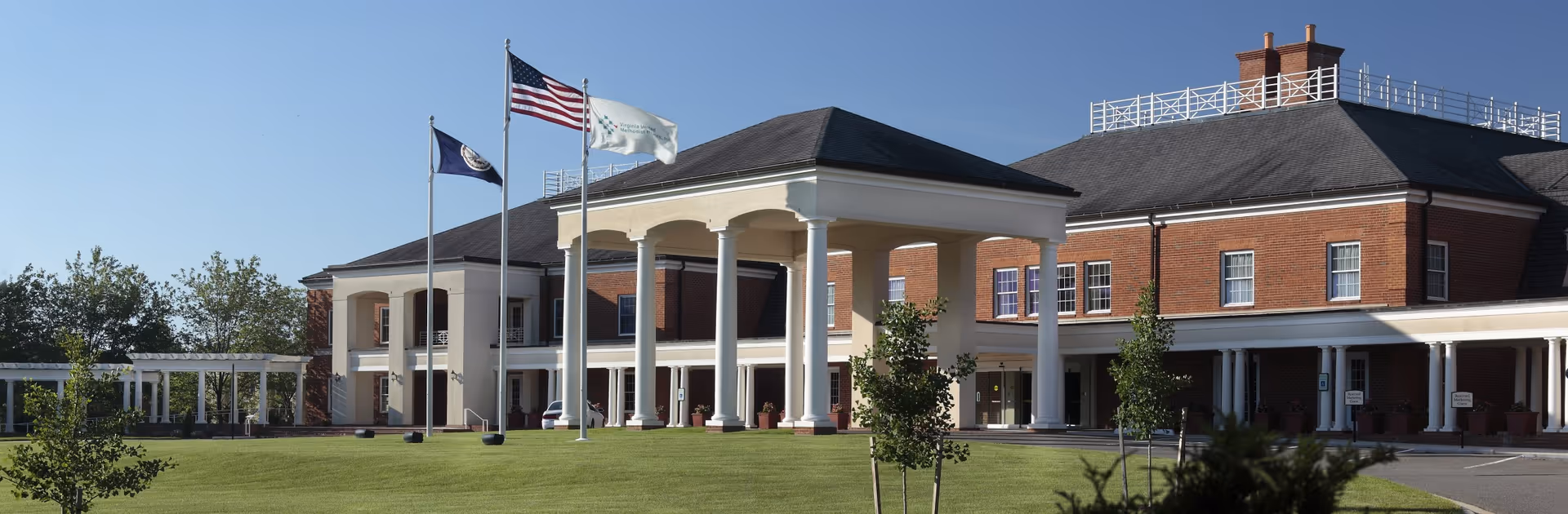 Front exterior view of a large brick building with white columns and a covered entrance. Three flagpoles with flags stand on a well-maintained lawn in front of the building under a clear blue sky.