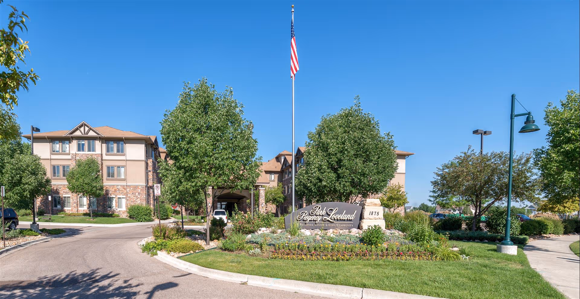 Front entrance of a multi-story senior living building with a landscaped circular driveway, flagpole, and entrance sign.