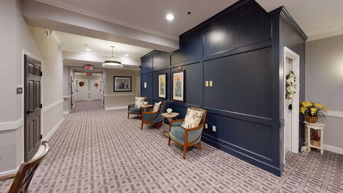 Carpeted hallway with a seating area and dark blue paneled accent wall featuring framed artwork inside a senior living facility.