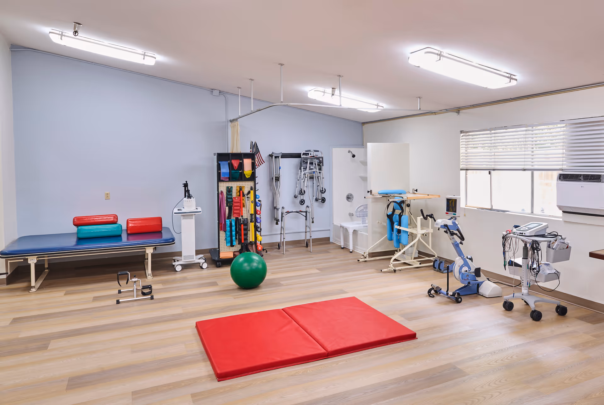A bright physical therapy room with various rehabilitation equipment including a treatment table with red and teal cushions, a green exercise ball, a red exercise mat on the wooden floor, a rack holding resistance bands and other therapy tools, walkers, a shower chair, and exercise machines near a window with blinds.