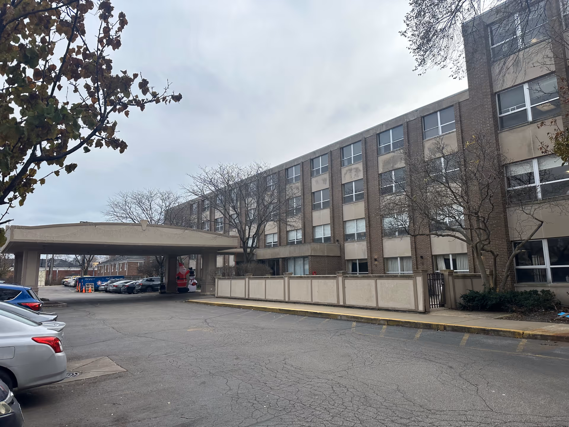 Exterior view of a multi-story senior living facility building with a covered entrance and a parking lot in front. There are several parked cars and some leafless trees around the building. Inflatable holiday decorations are visible under the covered entrance.