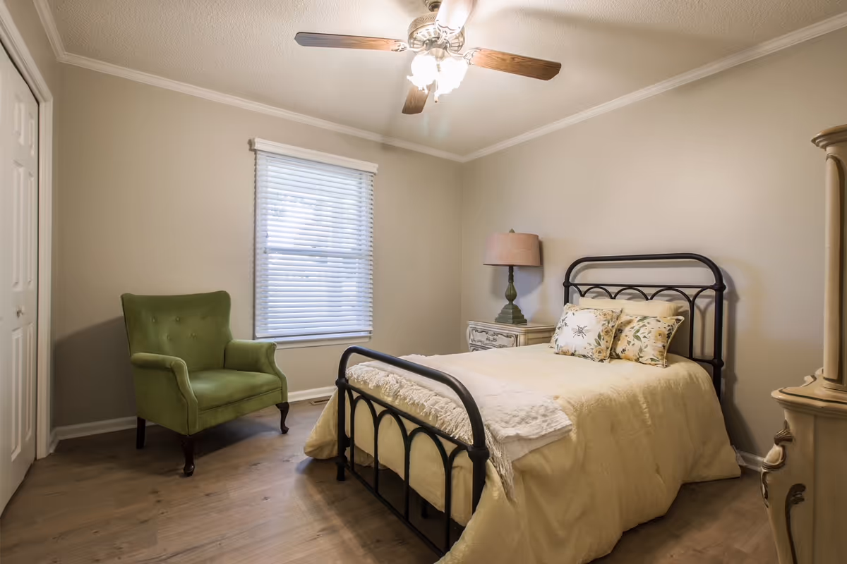 A cozy bedroom with a black metal bed frame, cream-colored bedding, and floral pillows. There is a green upholstered armchair near a window with white blinds, a vintage-style nightstand with a lamp, and wooden flooring. The walls are painted light beige and a ceiling fan with lights is on the ceiling.