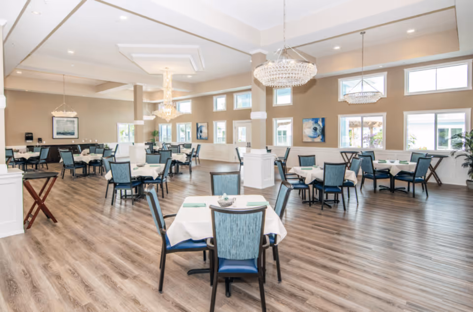 Bright, spacious dining room with multiple tables set with white linens, blue chairs, chandeliers, and large windows.