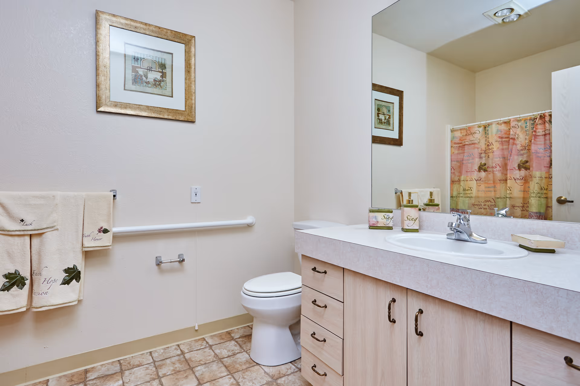 A clean bathroom with a toilet, a sink with countertop and cabinets, a large mirror, a towel rack with beige towels, a framed picture on the wall, and a shower curtain with a colorful pattern.