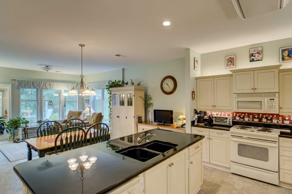 Bright and spacious kitchen and dining area in a senior living facility. The kitchen features white cabinets, a white stove, microwave, and a black countertop island with a sink. The dining area has a wooden table with six green chairs and a hanging light fixture above. In the background, there is a cozy sitting area with armchairs near large windows letting in natural light. The walls are decorated with framed artwork and a round clock.