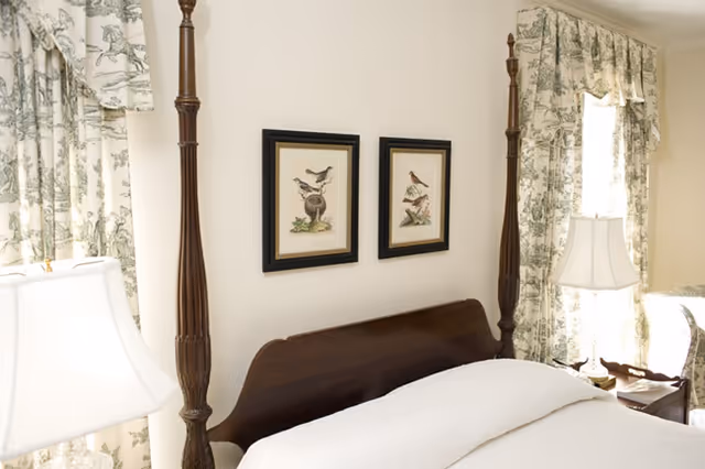 A bedroom with a wooden four-poster bed featuring white bedding. Two framed bird illustrations hang on the wall above the headboard. The room has floral-patterned curtains and a white table lamp on a bedside table.