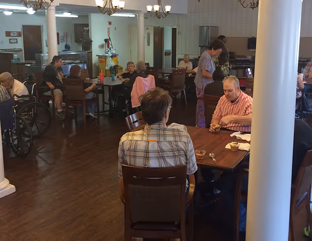 A dining area in a senior living facility with several elderly residents seated at tables eating and socializing. The room has wooden floors, chandeliers, and a kitchen area in the background. Some residents are in wheelchairs, and a caregiver is assisting one of them.