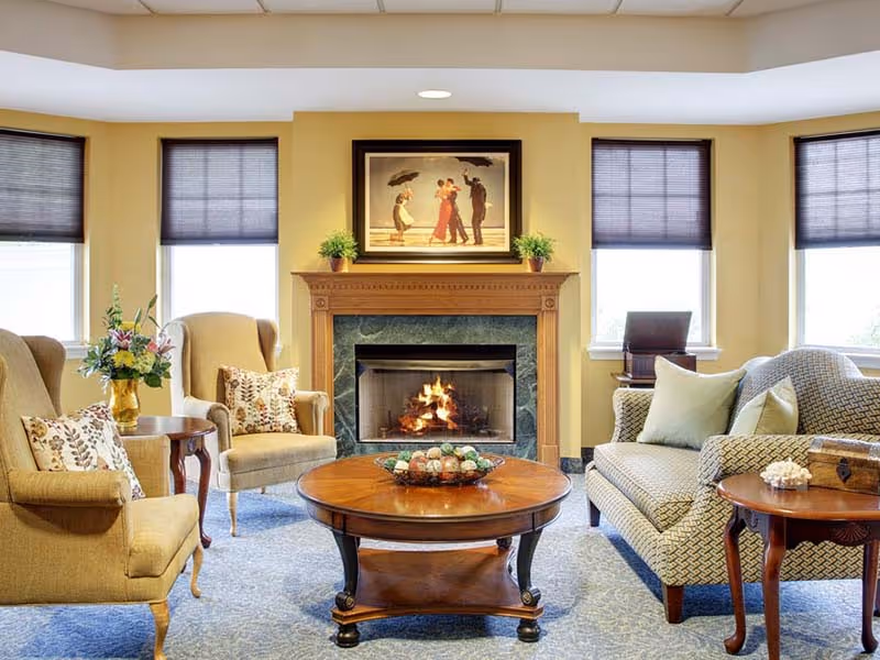 A cozy living room with a lit fireplace in the center, flanked by two potted plants on the mantel. Above the fireplace is a framed painting of people dancing in the rain. The room has two beige armchairs with floral pillows on the left, a patterned sofa with light-colored pillows on the right, and a round wooden coffee table with decorative items in the middle. There are three windows with dark blinds behind the seating area, and a side table with a flower vase next to one armchair.