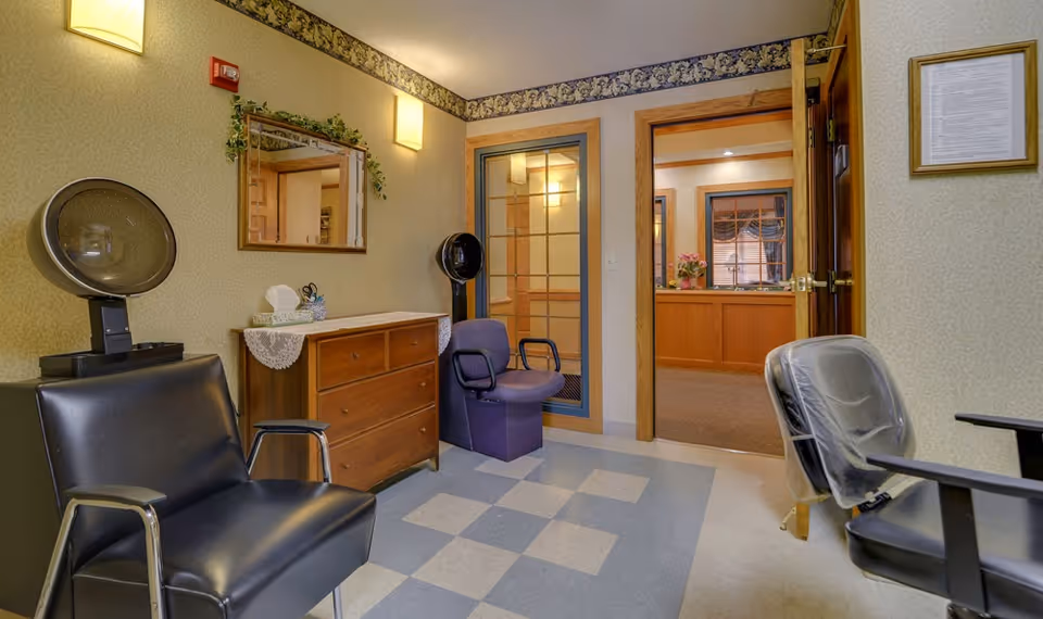 Interior view of a small salon area in a senior living facility with two black salon chairs, two hair dryers, a wooden dresser with a lace doily and tissue box, a wall mirror with greenery above it, and a doorway leading to another room with a wooden counter and window.