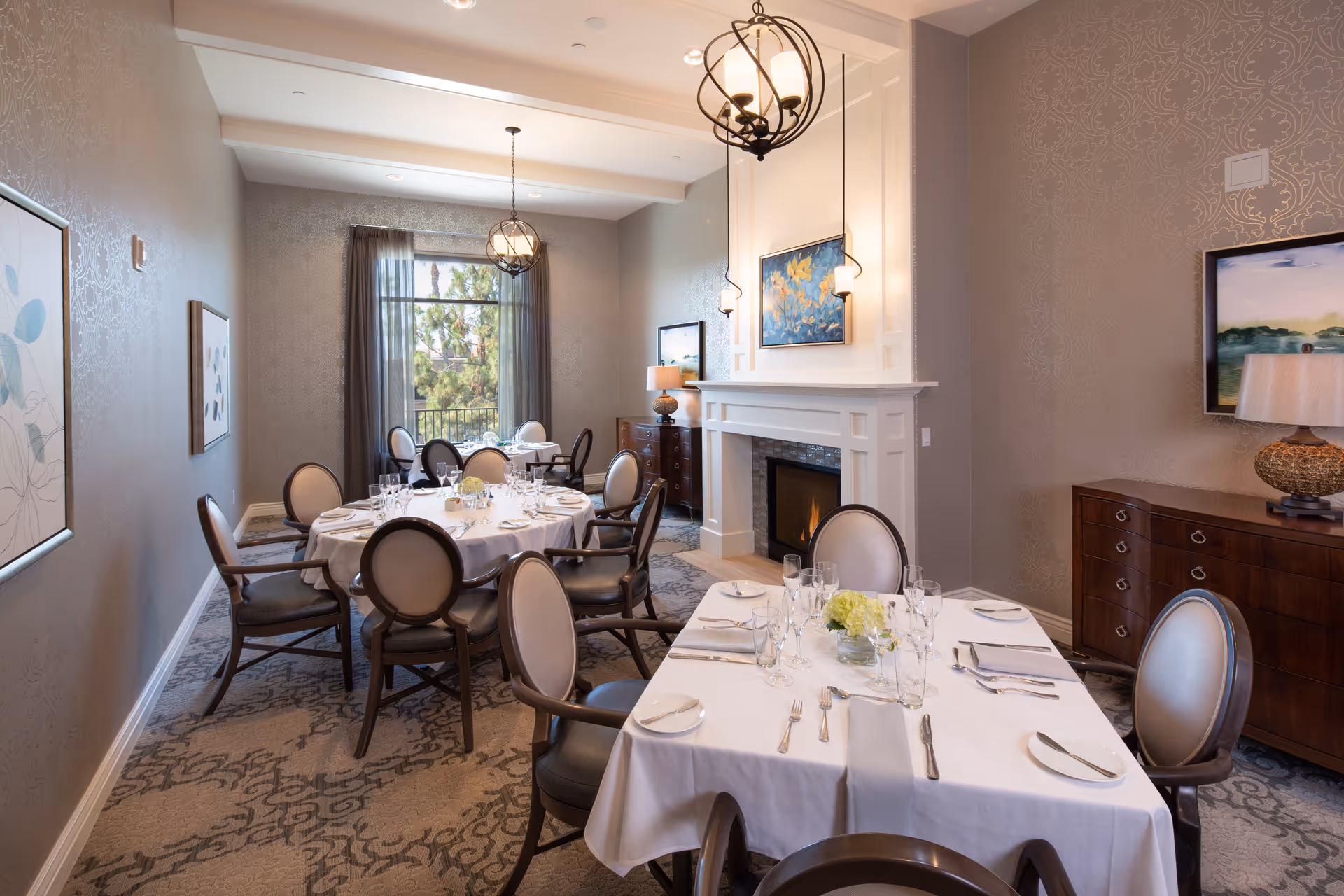 A well-lit dining room with round and rectangular tables set with white tablecloths, glassware, plates, and silverware. The room features elegant chairs with cushioned seats, a fireplace with a painting above it, two chandeliers hanging from the ceiling, and large windows with sheer curtains allowing natural light to enter. The walls are decorated with framed artwork and a sideboard with a lamp.