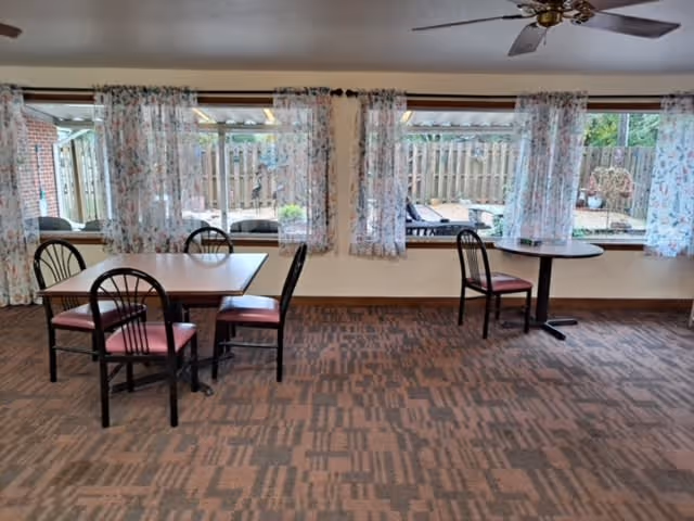 A dining area with two tables and several chairs with red cushions. Large windows with floral curtains look out onto a fenced outdoor patio area with lounge chairs and plants. The room has a patterned carpet and a ceiling fan.