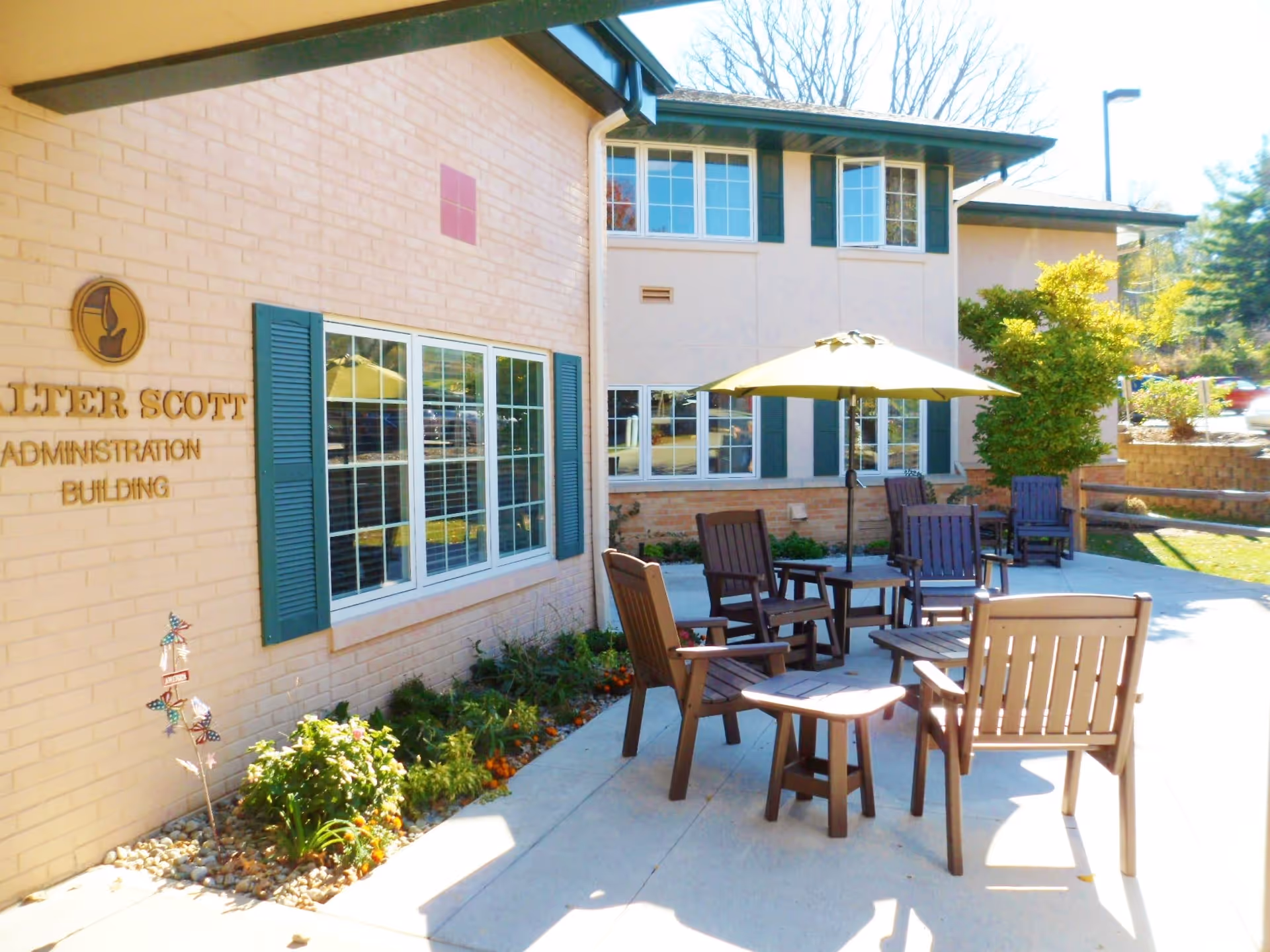 Outdoor patio area with several wooden chairs and tables, including one table with a yellow umbrella. The patio is adjacent to a beige building with green window shutters and a sign that reads 'Walter Scott Administration Building'. There are plants and flowers along the building's edge and a parking area visible in the background.