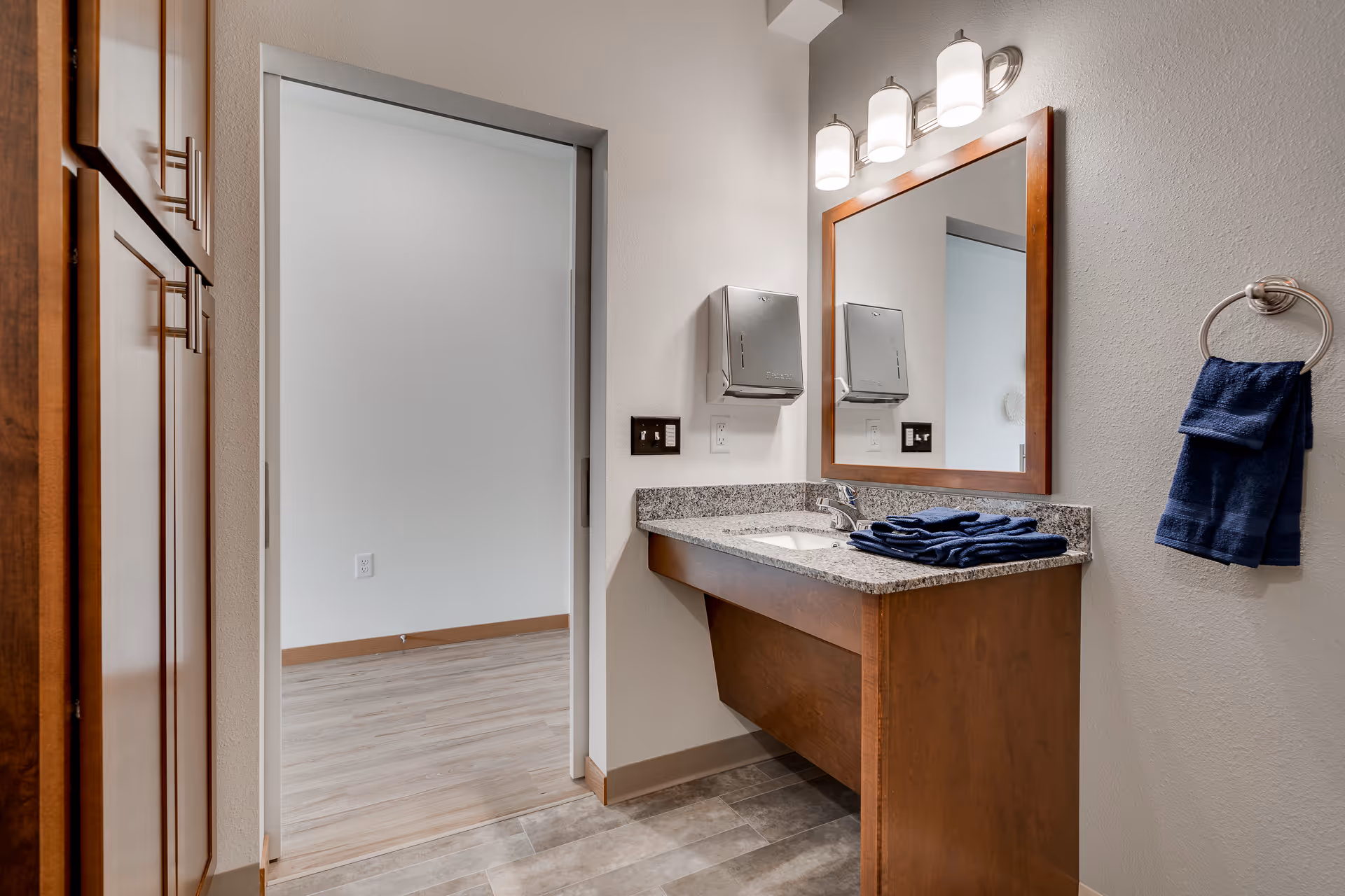A bathroom area with a wooden vanity topped with a granite countertop and a sink. Above the sink is a large mirror framed in wood, with three light fixtures mounted above it. Two stainless steel paper towel dispensers are mounted on the wall next to the mirror. A blue towel is hanging on a silver towel ring on the right wall, and additional blue towels are folded on the countertop. To the left, there is a wooden cabinet and an open doorway leading to a room with light wood flooring and white walls.