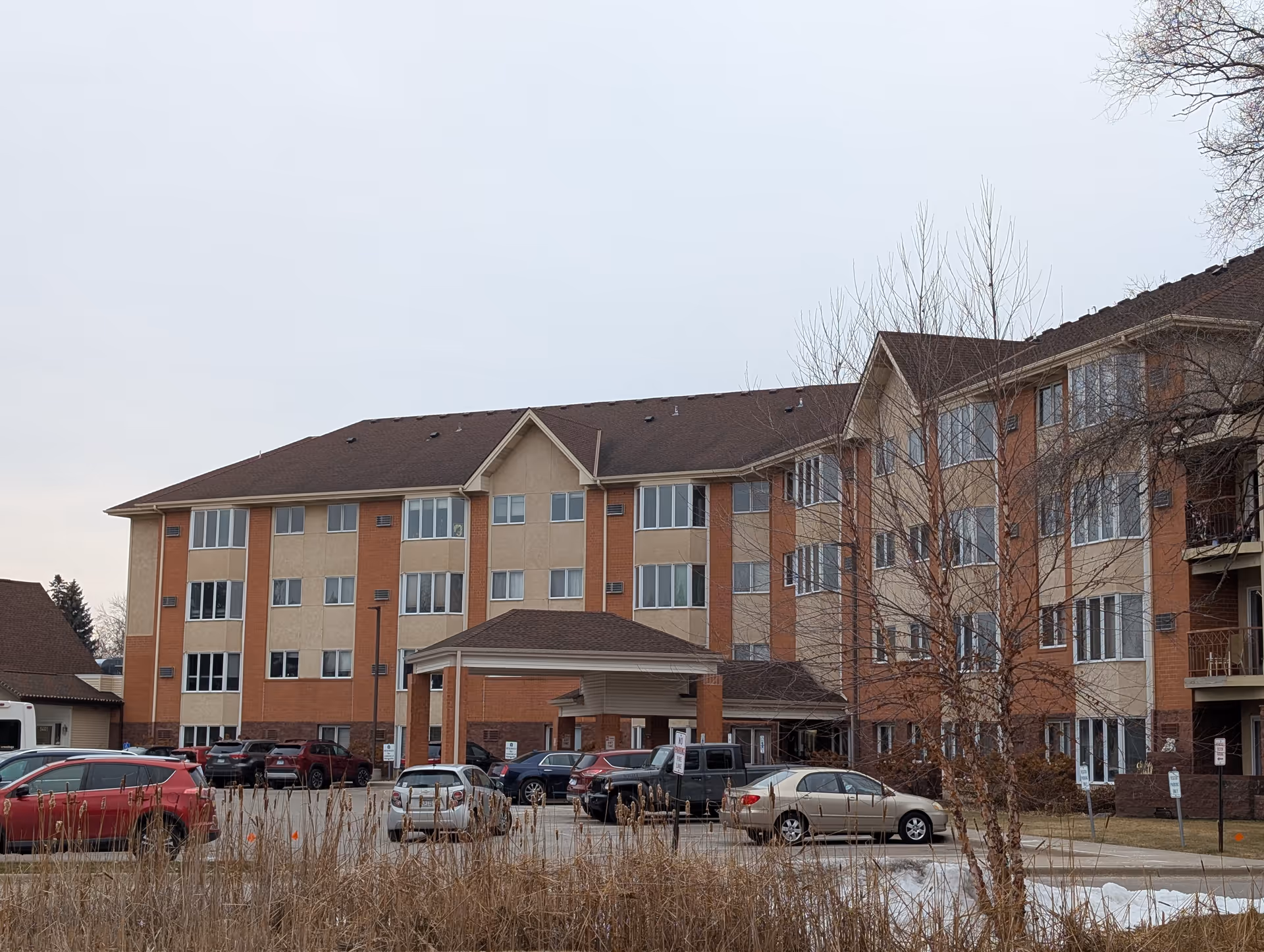 Exterior view of a multi-story senior living facility building with a covered entrance and several parked cars in front. The building has a beige and brick facade with many windows and leafless trees in the foreground.