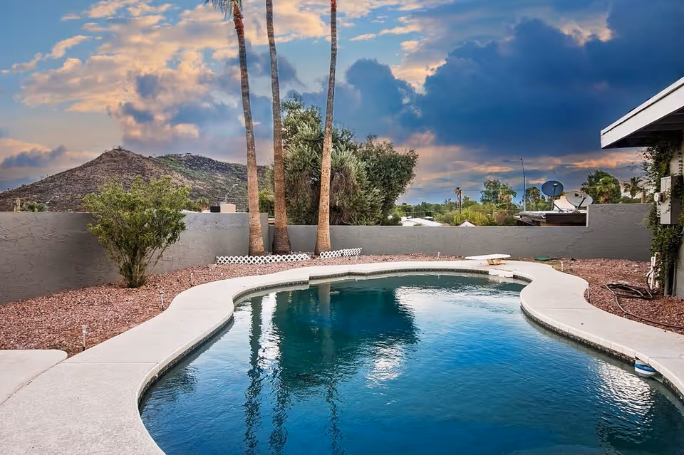 Outdoor swimming pool with a curved edge surrounded by a concrete deck and landscaping with rocks and plants. In the background, there are three tall palm trees, a gray wall, and a mountain under a partly cloudy sky during sunset.