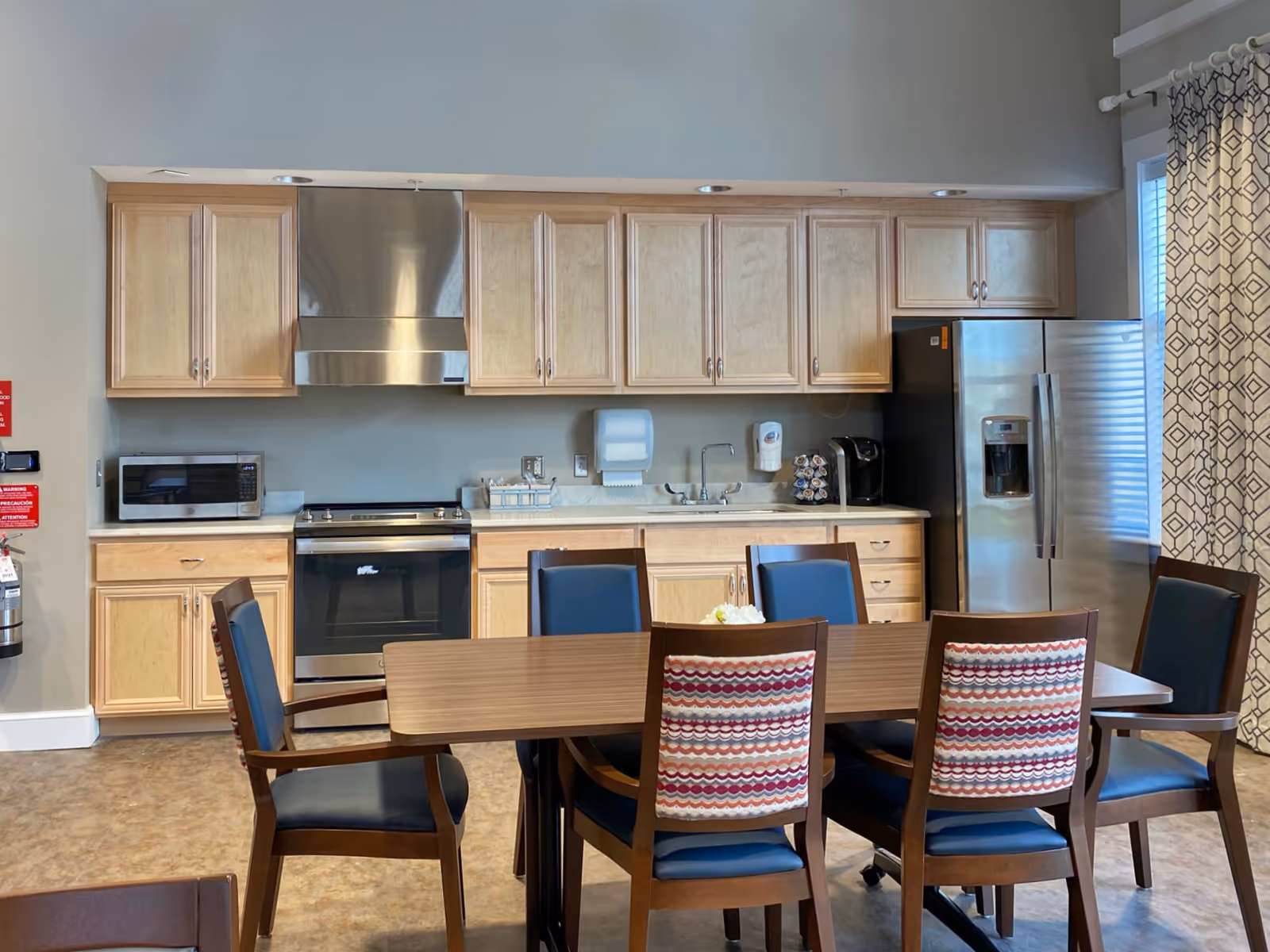 A kitchen area with light wood cabinets, a stainless steel stove and range hood, a microwave, a sink, a coffee maker, and a stainless steel refrigerator. In front of the kitchen is a wooden dining table with six chairs, two of which have patterned backs. A window with patterned curtains is visible on the right side.