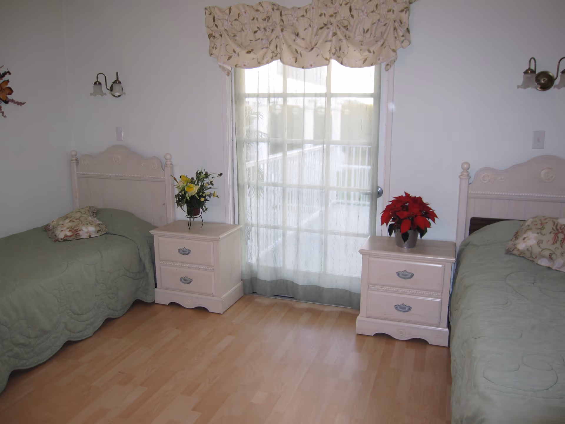 A bedroom with two twin beds, each with a green quilt and decorative pillow. Between the beds are two matching nightstands, each with a flower arrangement on top. A large window with sheer curtains and a floral valance is centered on the wall behind the nightstands. The room has light-colored wooden flooring and white walls.